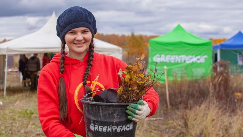 Ein Mädchen mit langen Zöpfen hält einen Eimer mit Setzlingen, im Hintergrund ist ein grünes Greenpeace Zelt zu sehen.